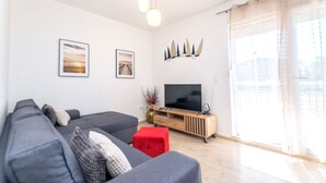 A modern living room with a grey sofa, a red ottoman, a wooden TV stand, and a large window with sheer curtains. - Apartment Beleca (Podstrana)