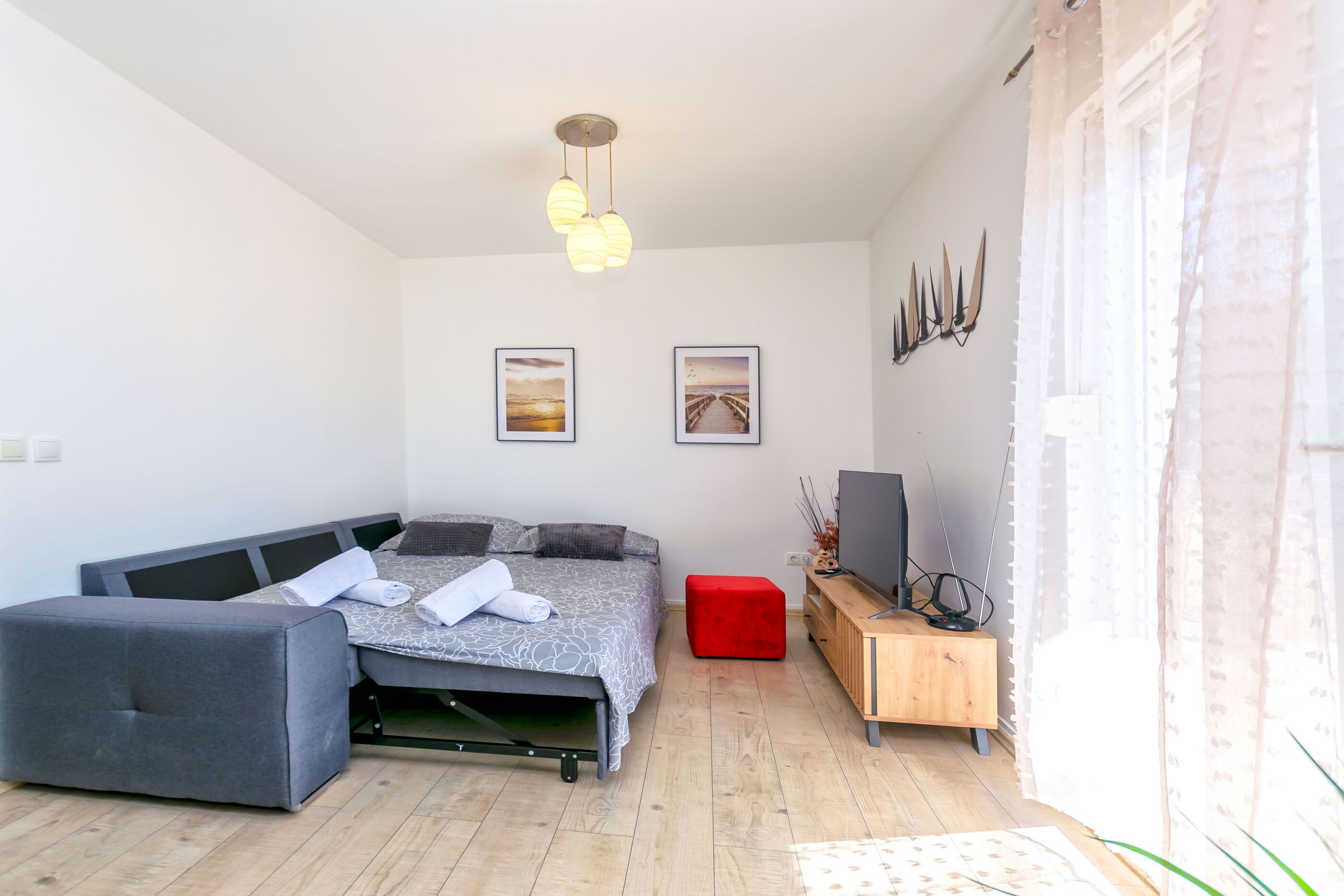 Une chambre à coucher moderne avec un lit, un pouf rouge, une télévision et une armoire en bois.