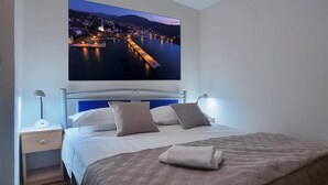 A neatly made bed with a large cityscape photo above it, a bedside table with a lamp, and a nightstand with a drawer. - House Zlatni Otok (Sibenik)