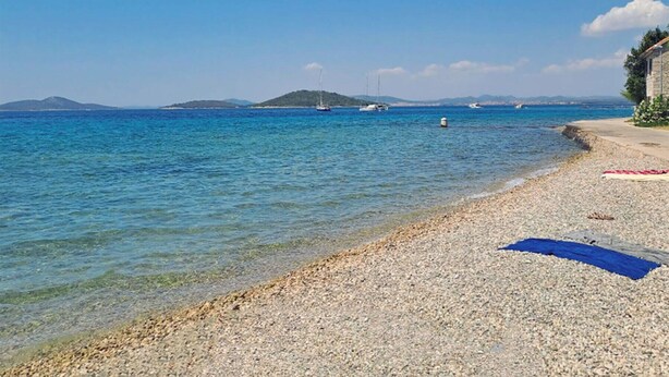 A pebble beach with clear blue water, boats, and distant hills. - House Zlatni Otok (Sibenik)