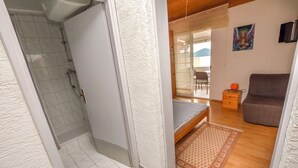 A bathroom with a shower, a toilet, and a sink. There is a bed with a patterned rug, a chair, and a small table with a lamp. - Apartment Mladen (Pakostane)
