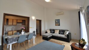 Een moderne woonkamer met een bank, een eettafel en stoelen. Er is een keuken met kastjes en een spoelbak.
