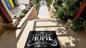 A black doormat with the word "HOME" in white letters, a stone pathway leading to a gate, and a row of green and yellow recycling bins to the left.