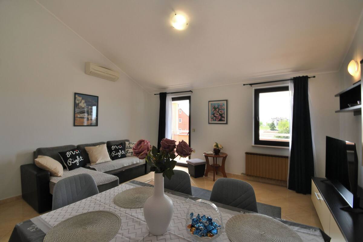 A modern living room with a sofa, a dining table with placemats, a vase with flowers, and a television.