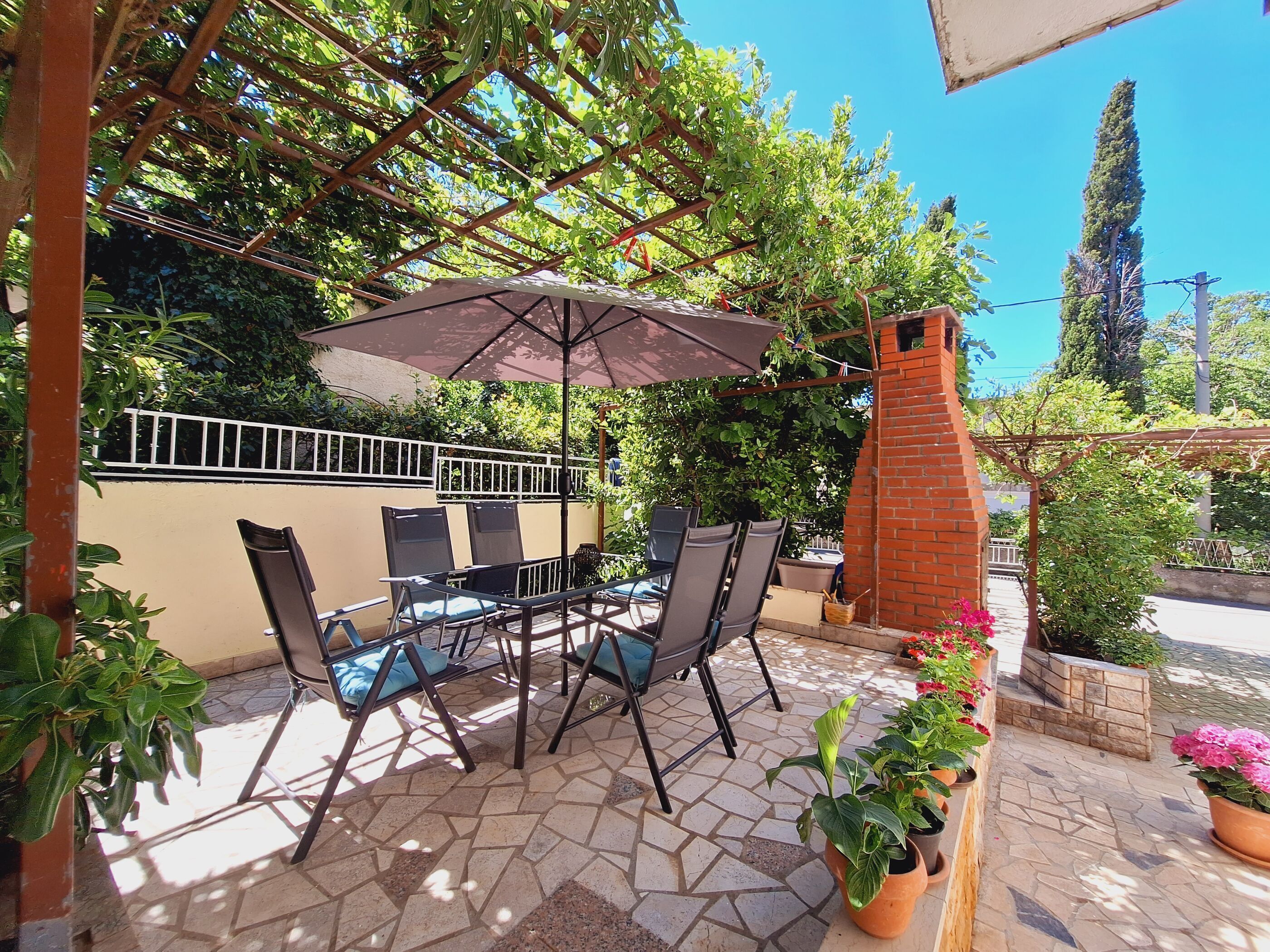 Un patio con tavolo e sedie sotto una pergola, circondato da piante in vaso e una struttura in mattoni.