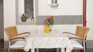 Une table à manger recouverte d’une nappe en marbre blanc, deux chaises en osier, un vase rempli de fleurs et une carafe en verre contenant du jus d’orange.