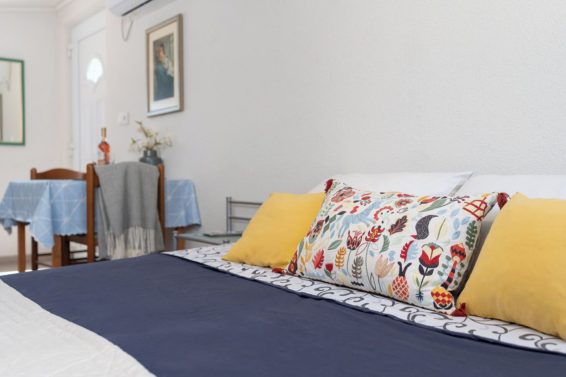 Ein ordentlich bezogenes Bett mit einem gemusterten und zwei einfarbigen Kissen, ein kleiner Tisch mit blauer Tischdecke und ein Stuhl.