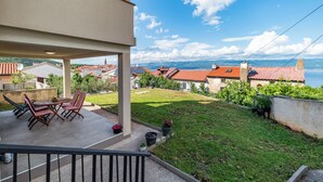 A balcony with a table and chairs, a grassy yard, and a view of buildings and a body of water. - Apartment Krtica (Vrbnik)