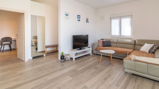 A modern living room with a flat-screen TV on a white stand, a beige sofa, a small round table, and a large window. - Apartment Krtica (Vrbnik)