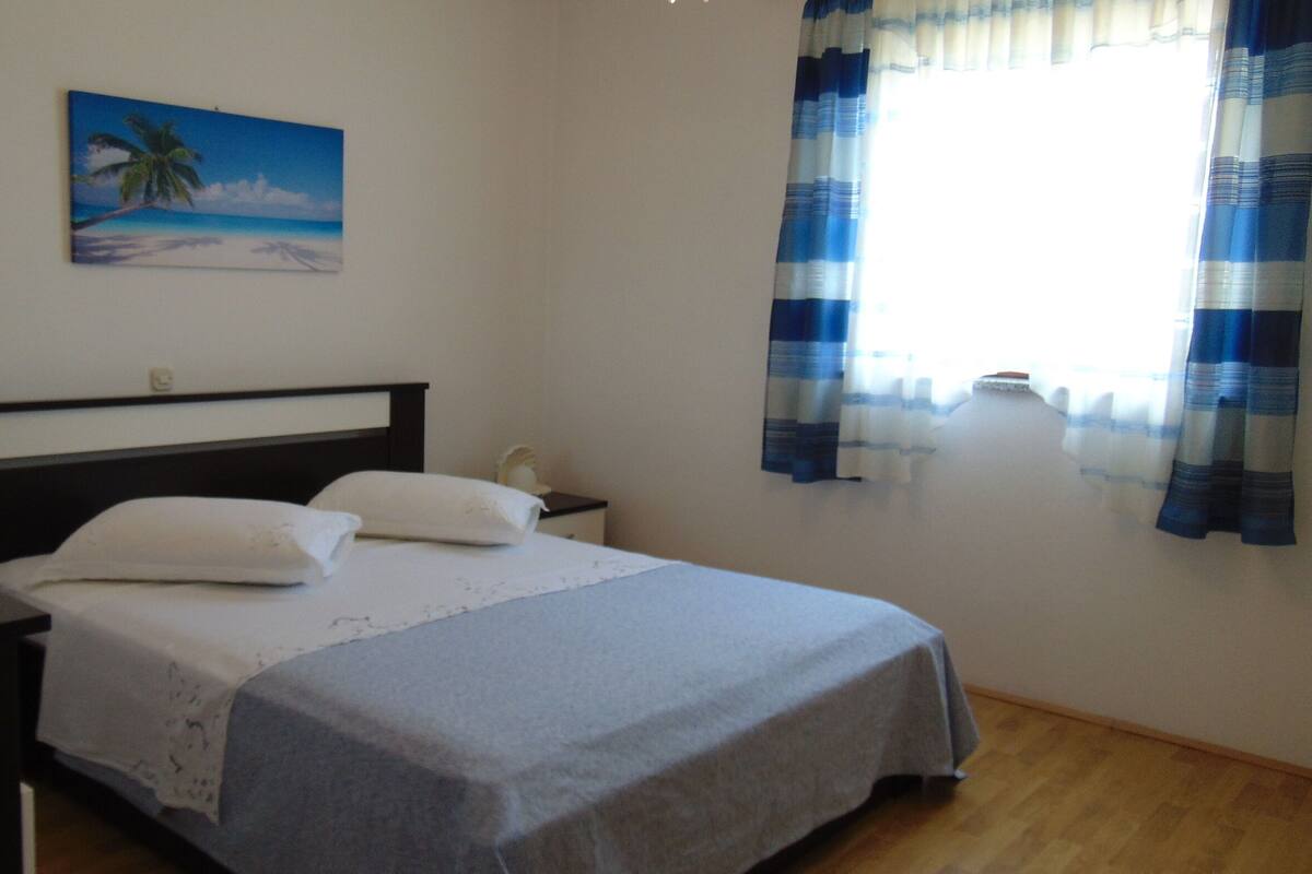 A bedroom with a bed, a nightstand, a painting of a beach scene, and a window with curtains.