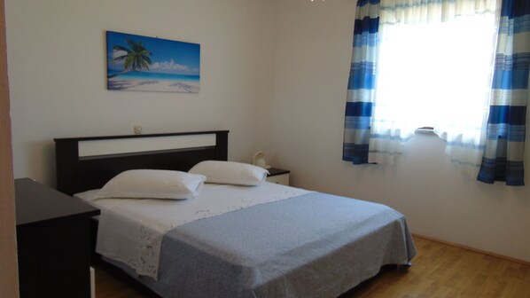 A bedroom with a bed, a nightstand, a painting of a beach scene, and a window with curtains. - Apartment Ana (Jasenice)
