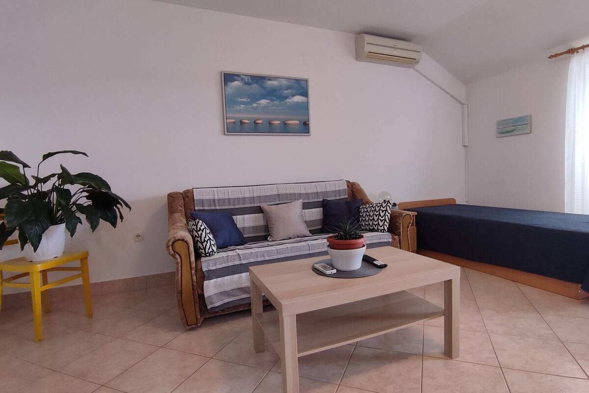 A room with a sofa, a bed, a small table, a potted plant, and a painting on the wall.