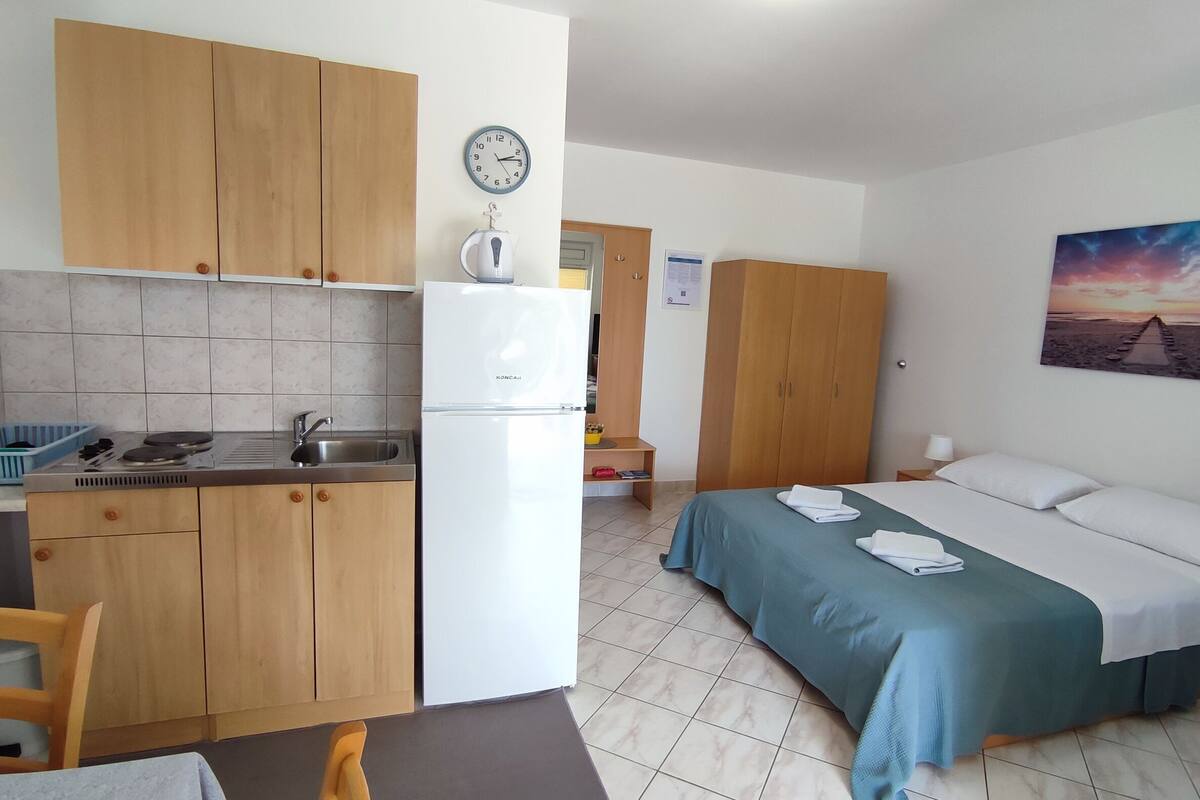 A compact hotel room with a kitchenette, a bed with a view of the beach, and a painting on the wall.