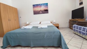 Un lit bien fait, recouvert d’une couvre-lit turquoise et agrémenté de serviettes blanches, une armoire en bois, un tableau représentant une scène de plage, un téléviseur posé sur un meuble en bois et un sol carrelé.
