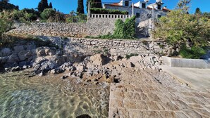Un muro de piedra con una piscina cristalina al frente, una escalera de piedra y un edificio con antenas parabólicas al fondo.