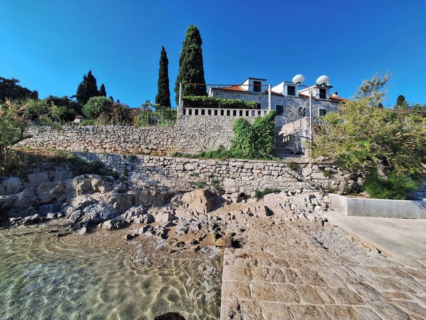 A stone retaining wall with a clear water pool in front, a stone staircase, and a building with satellite dishes in the background. - Apartments Neptun (Solta)