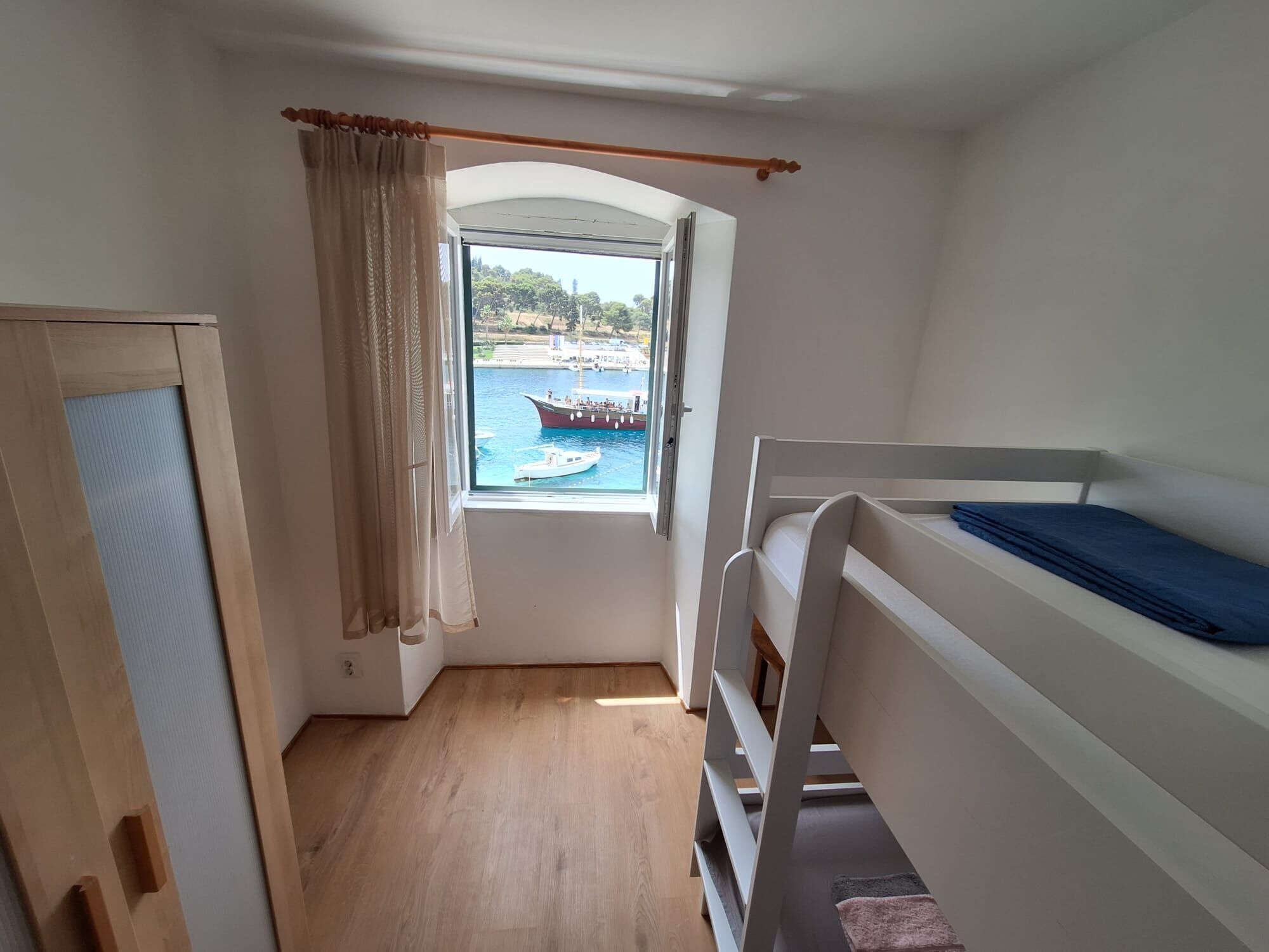 A room with a bunk bed, a window with a view of boats on water, and a door with a curtain.