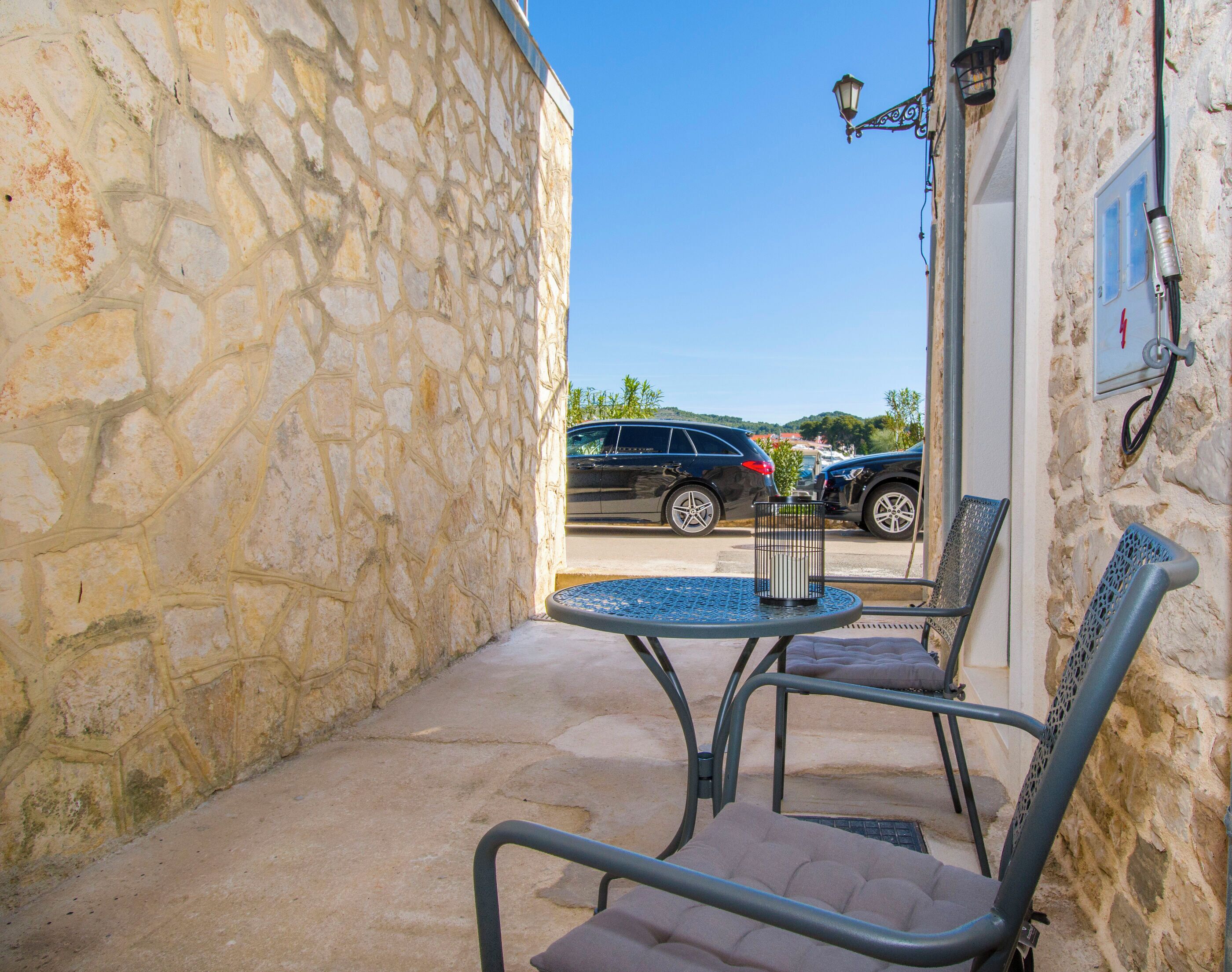 A stone-walled outdoor seating area with metal chairs and a small round table, overlooking parked cars and a clear blue sky.