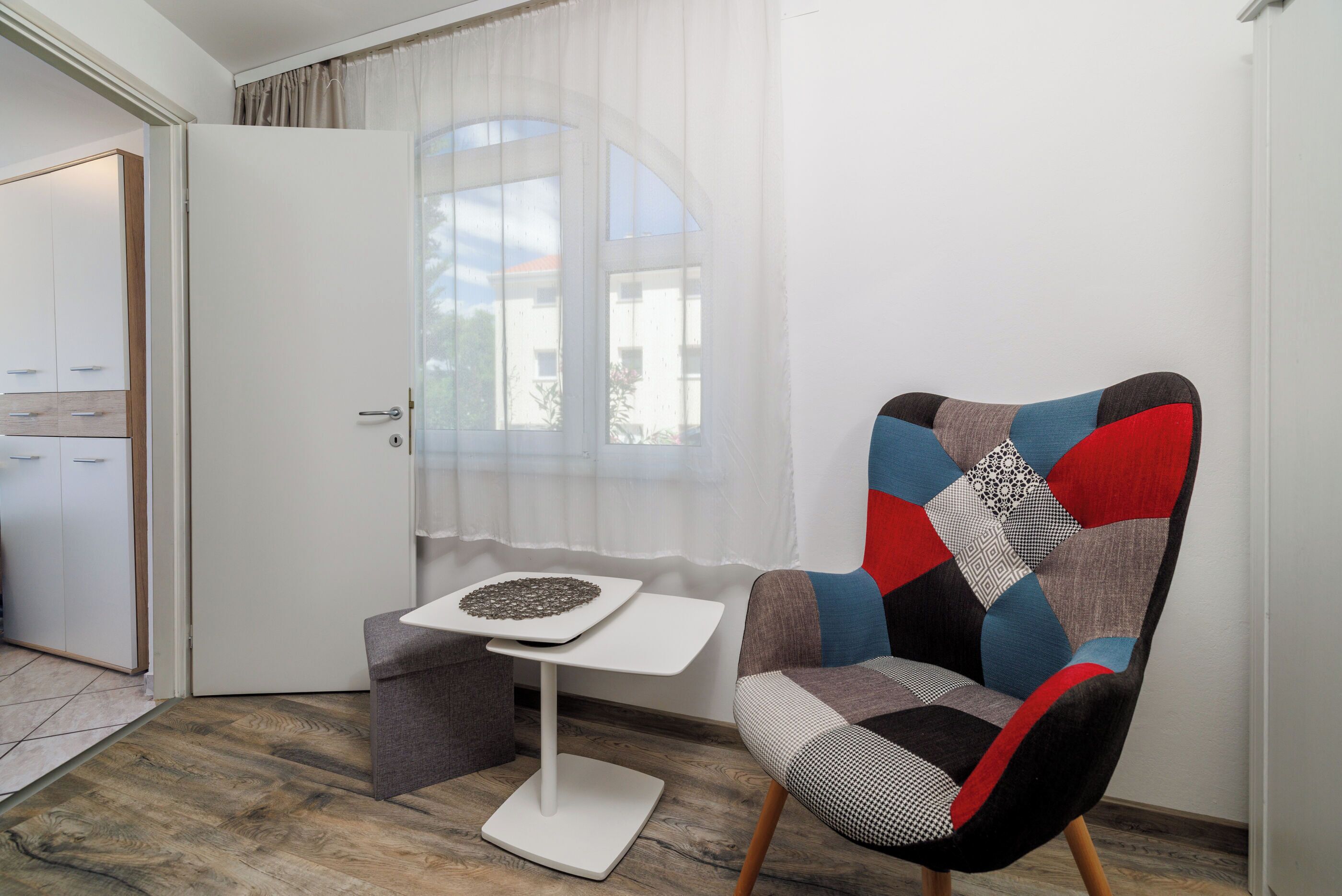 A room with a wooden floor, a white door, a window with sheer curtains, a small white table with a decorative object, and a colorful armchair.