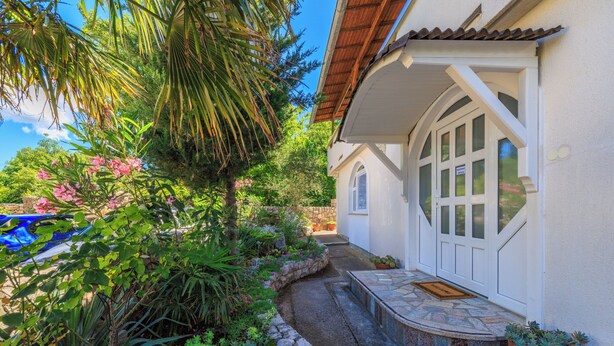 A house with a stone pathway leading to a white door, surrounded by lush greenery and a blue car parked nearby. - Apartment Lokvica (Omisalj)