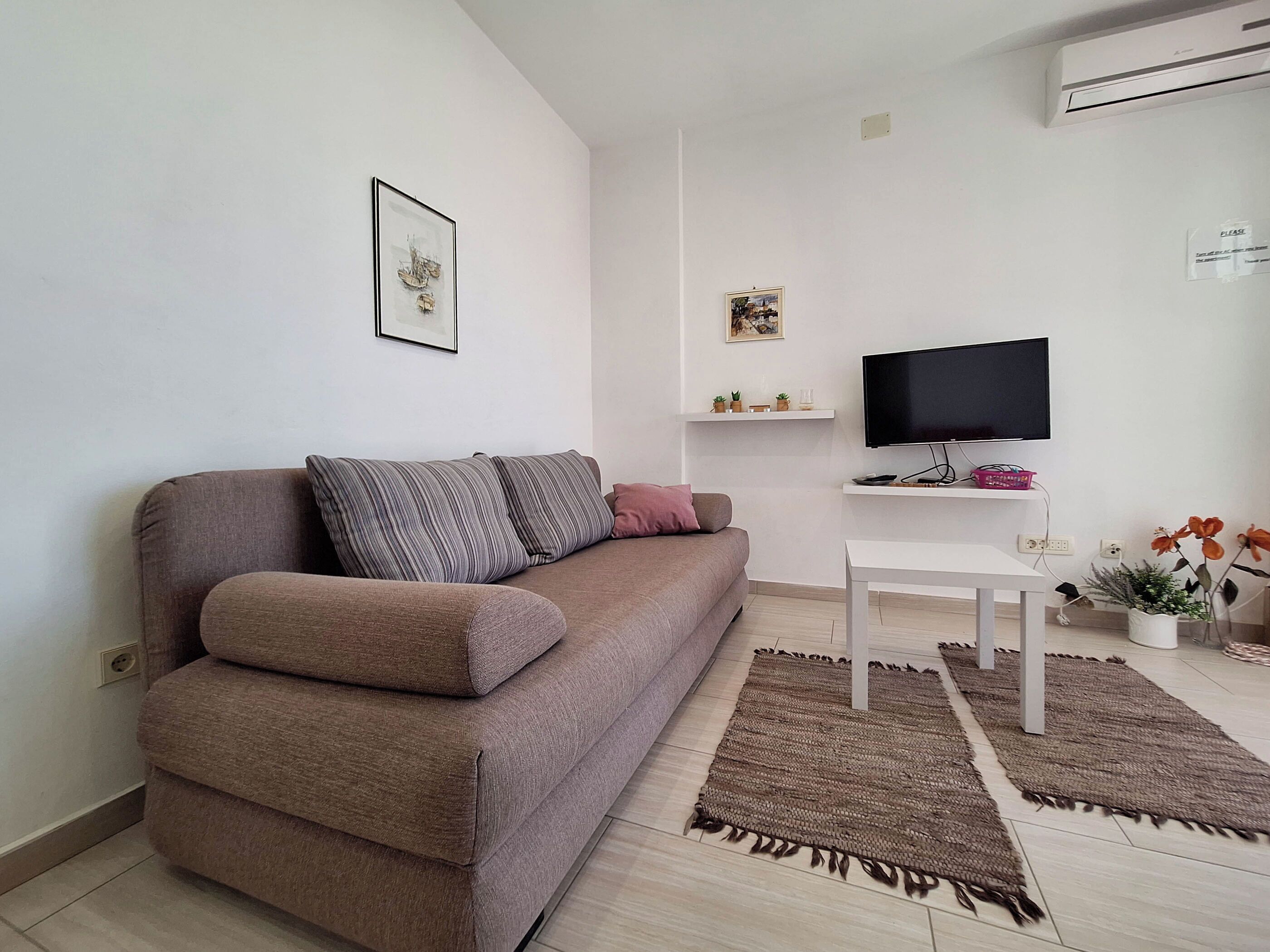 A modern living room with a brown sofa, a white coffee table, a flat-screen TV, and decorative items on shelves.