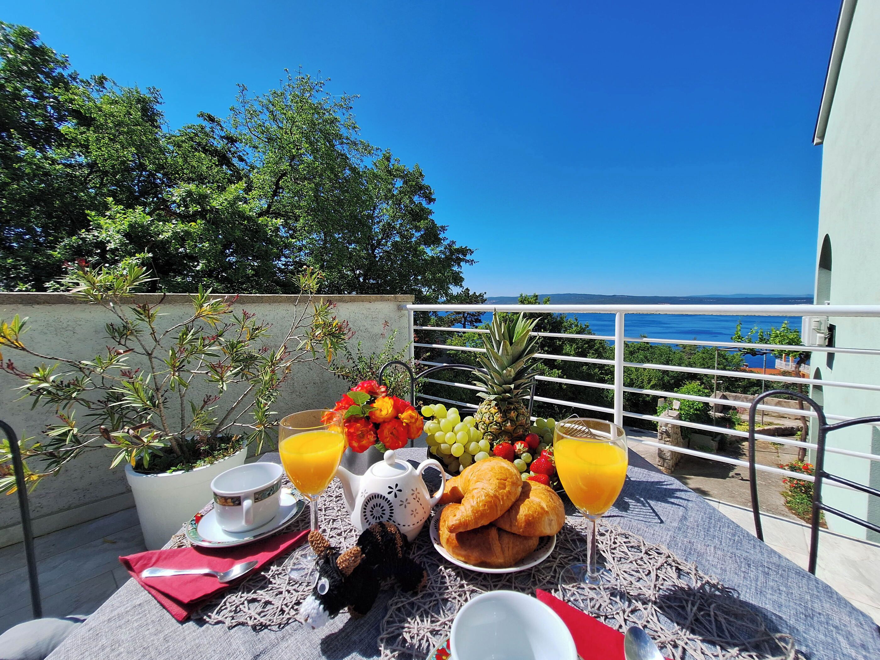 A balcony with a table set for breakfast, including croissants, fruit, and juice, overlooking a scenic coastal view.
