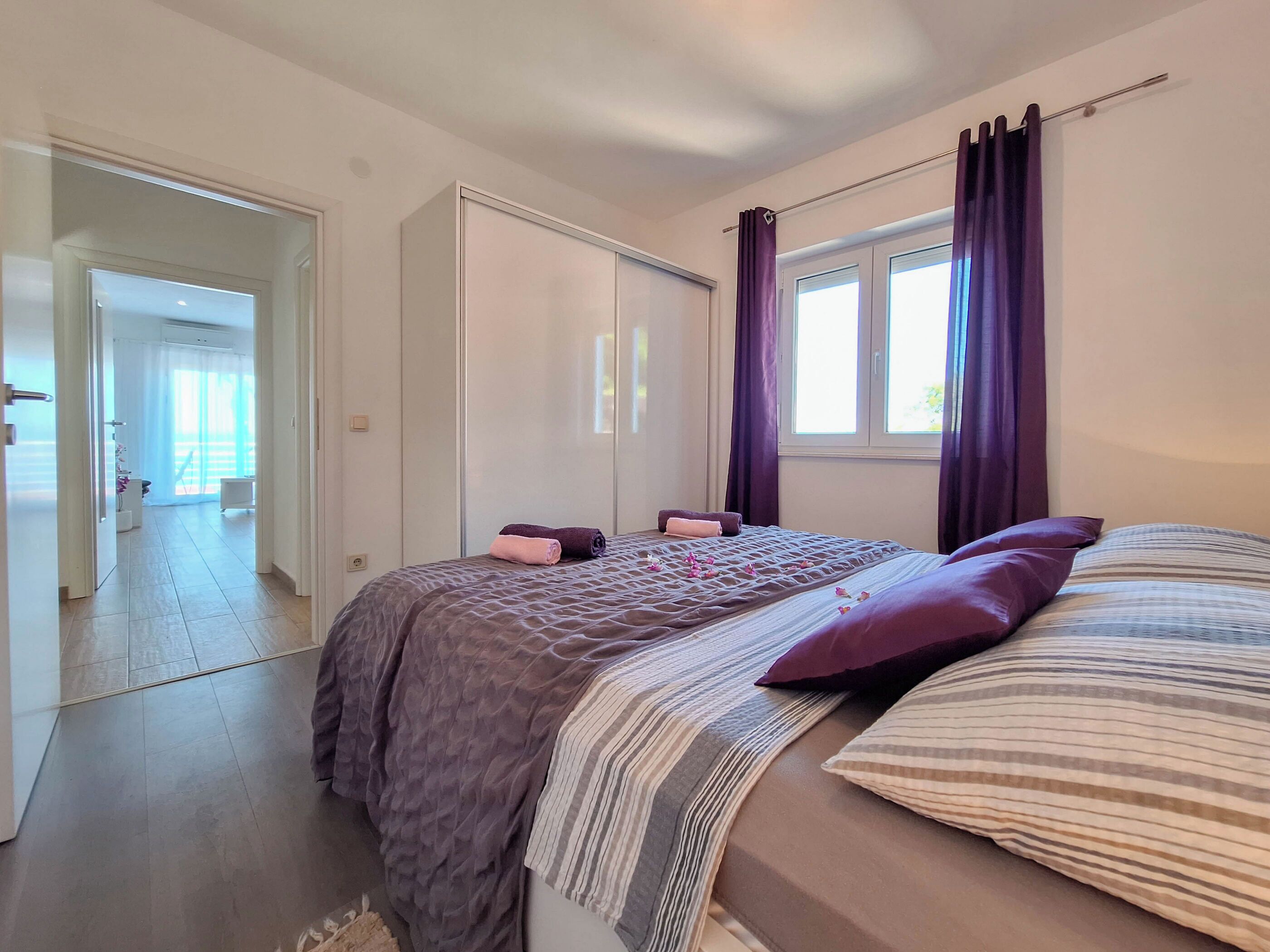 Um quarto com cama, janela com cortinas, porta e corredor que dá para outro espaço.