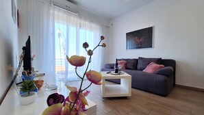 Een moderne woonkamer met een grijze bank, een witte salontafel en een groot raam met transparante gordijnen.