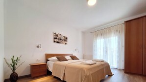 A bedroom with a wooden bed, a nightstand, a bedside lamp, a potted plant, and a large window with sheer curtains. - Apartment and Rooms Katunar (Crikvenica)