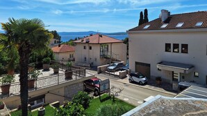 A residential area with a view of the sea, featuring houses with tiled roofs and balconies, a palm tree, and parked cars. - Apartment and Rooms Katunar (Crikvenica)