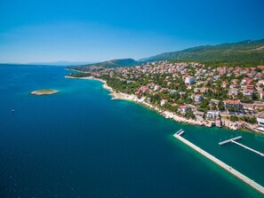 An aerial view of a coastal town with a pier extending into the sea, surrounded by residential buildings and greenery. - Apartments Panos (Novi Vinodolski)