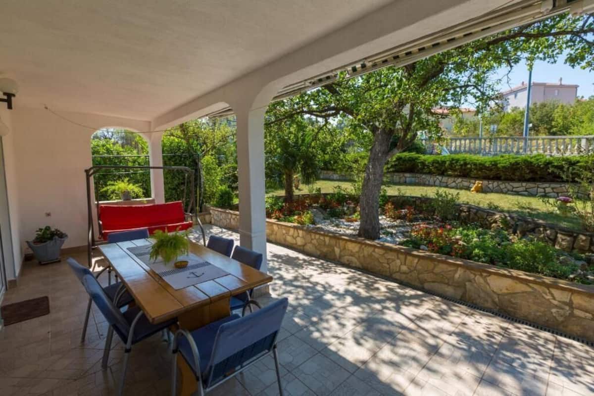 An outdoor patio with a wooden table and chairs, a red bench, and a view of a garden with trees and a stone wall.