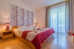 A hotel room with a large bed, two bedside lamps, a wall art with leaves, and a view of greenery through the window. - Apartments Panos (Novi Vinodolski)