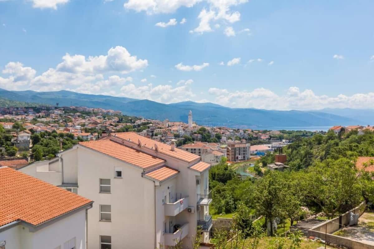 A coastal town with terracotta-roofed buildings, a prominent tower, and a view of the sea and mountains.