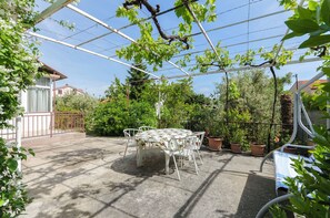 Eine überdachte Terrasse mit Tisch und Stühlen, umgeben von Topfpflanzen und einer Pergola.