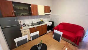 A compact kitchen with a refrigerator, stove, sink, and wooden cabinets. A red sofa is positioned near the window. - Apartments Luna (Marina)