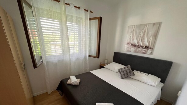 A bedroom with a bed, a nightstand, a window with curtains, a painting on the wall, and a door. - Apartments Luna (Marina)