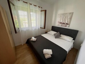 A bedroom with a bed, a nightstand, a window with curtains, a painting on the wall, and a door. - Apartments Luna (Marina)