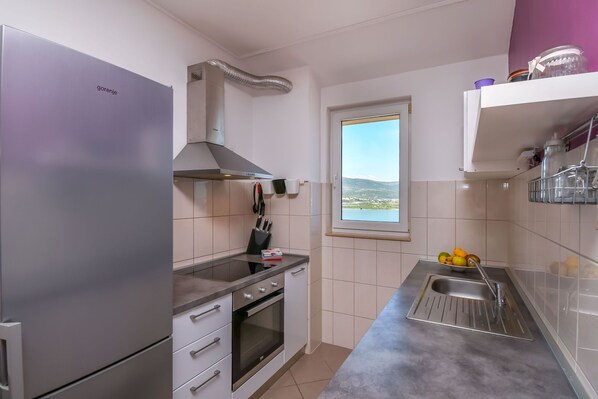 A compact kitchen with a stainless steel refrigerator, oven, and sink. A window provides a view of the outdoors. - Apartment Dani (Trogir)