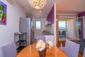 Eine moderne Küche mit Kühlschrank, Backofen und Spüle. Ein Esstisch mit Stühlen und eine Blumenvase sind vorhanden. Durch die Schiebetüren ist ein Balkon mit Bergblick zu sehen.
