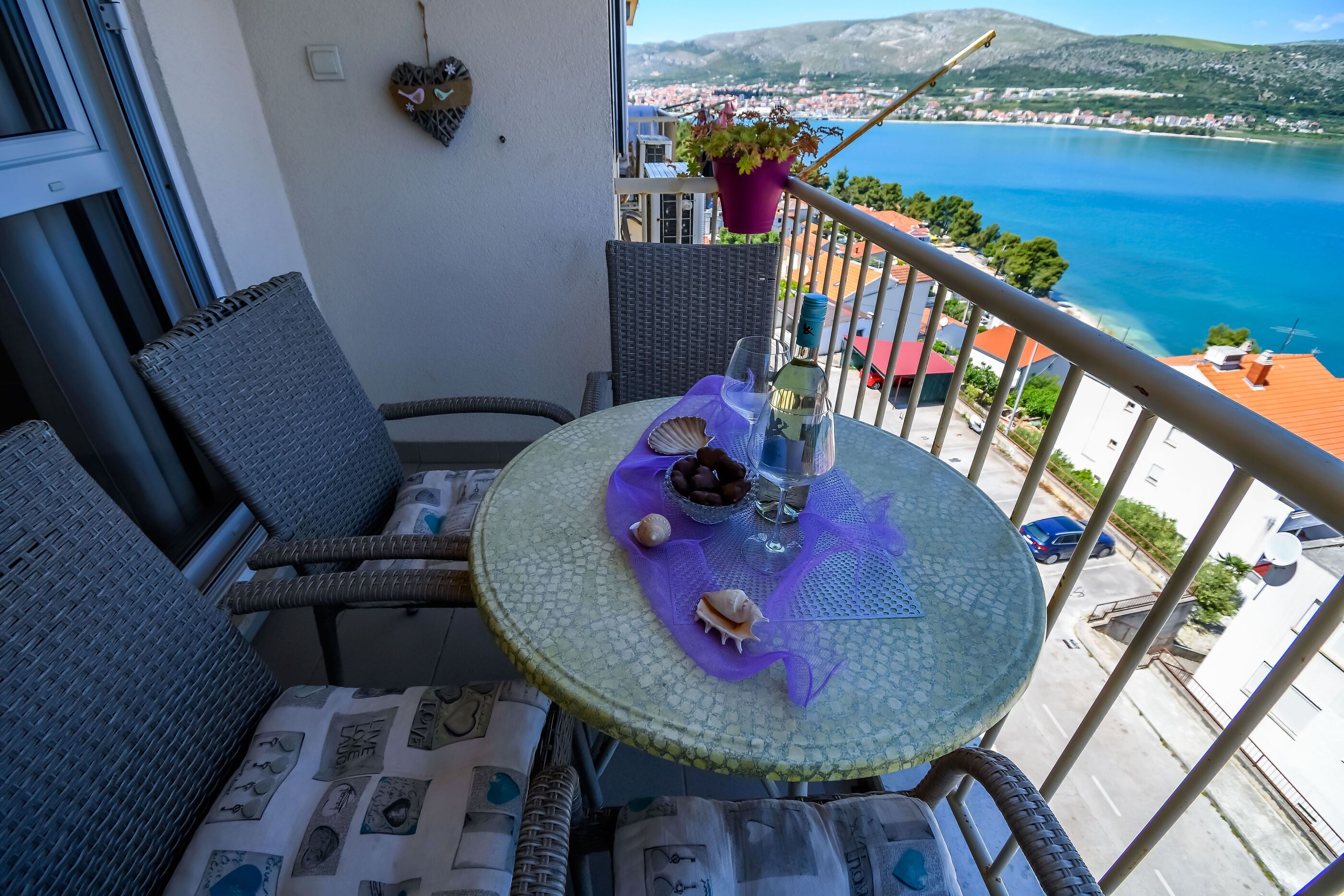 A balcony with wicker furniture, a table with decorative items, and a view of a coastal town and sea.