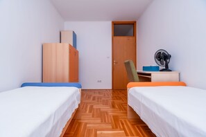 A small, well-lit room with two single beds, a wooden wardrobe, a desk with a chair, and a standing fan. - Apartment Dani (Trogir)