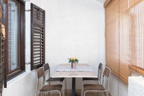 Un petit patio clos, aménagé avec une table et quatre chaises, un vase rempli de fleurs jaunes et une fenêtre ornée de volets en bois.