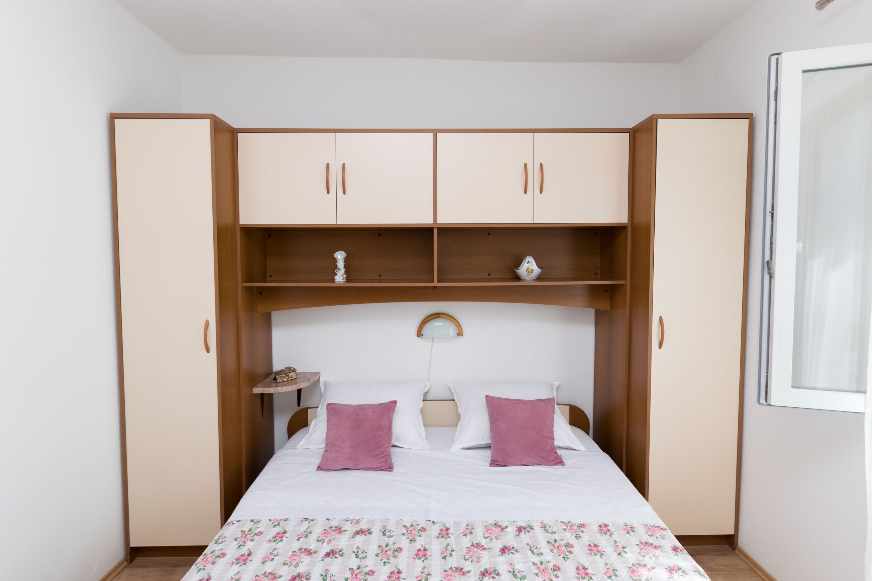 A bedroom with a bed, two pink pillows, a wooden wardrobe, and a small table with a lamp.
