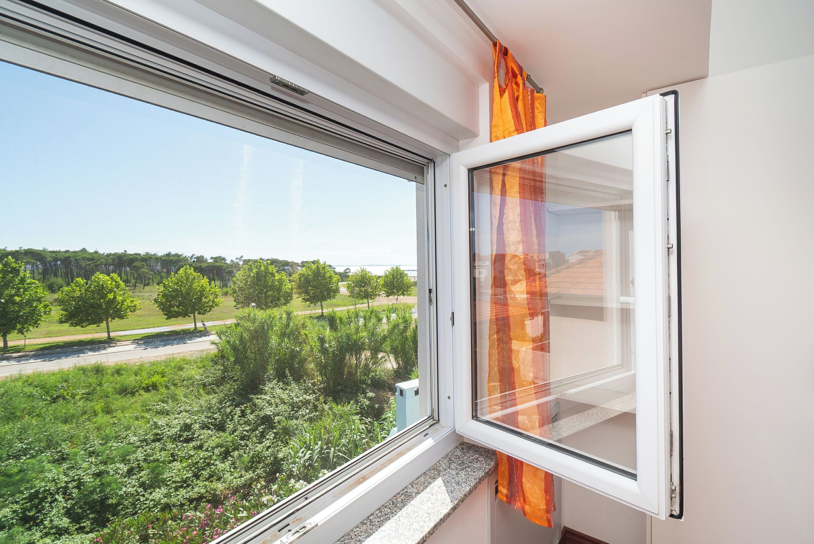 A window with a view of a green landscape, a white wall, and a curtain.