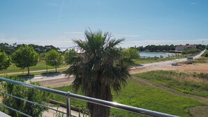 A view from a balcony overlooking a road with a palm tree in the foreground, a body of water, and buildings in the distance. - Apartment Terezija by the sea (Privlaka)