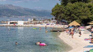 Ein Strand mit badenden und sonnenden Menschen, einer Promenade und Bergen im Hintergrund.