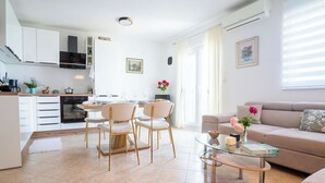 Uma cozinha moderna com armários brancos, um espaço de jantar com mesa de vidro e uma sala de estar com sofá e mesinha de centro.