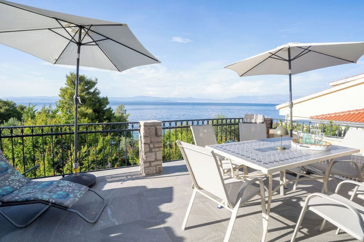 Ein Balkon mit Tisch, Stühlen und zwei Sonnenschirmen, mit Blick aufs Meer.