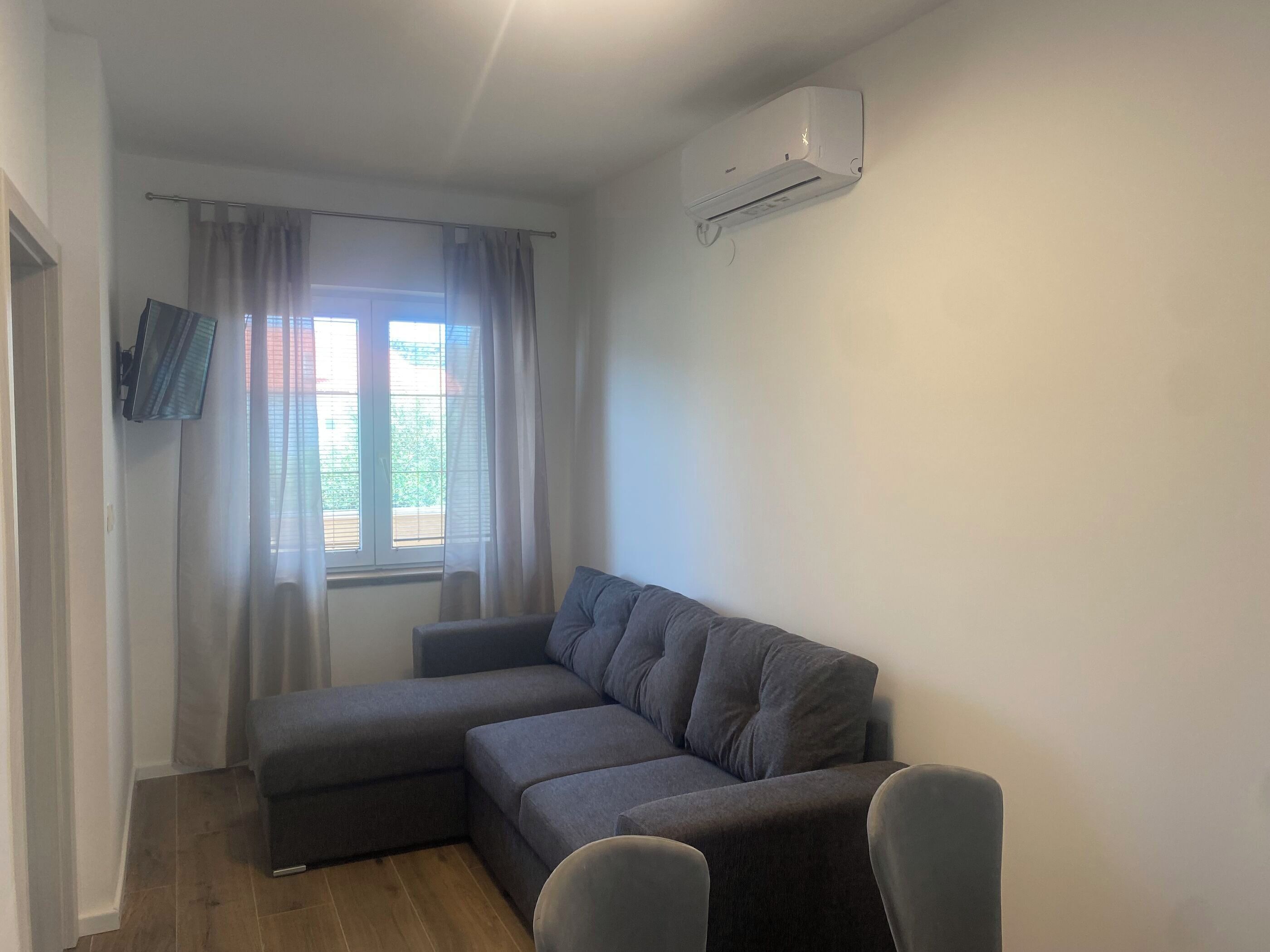 A modern living room with a grey sectional sofa, a flat-screen TV mounted on the wall, and a window with sheer curtains.